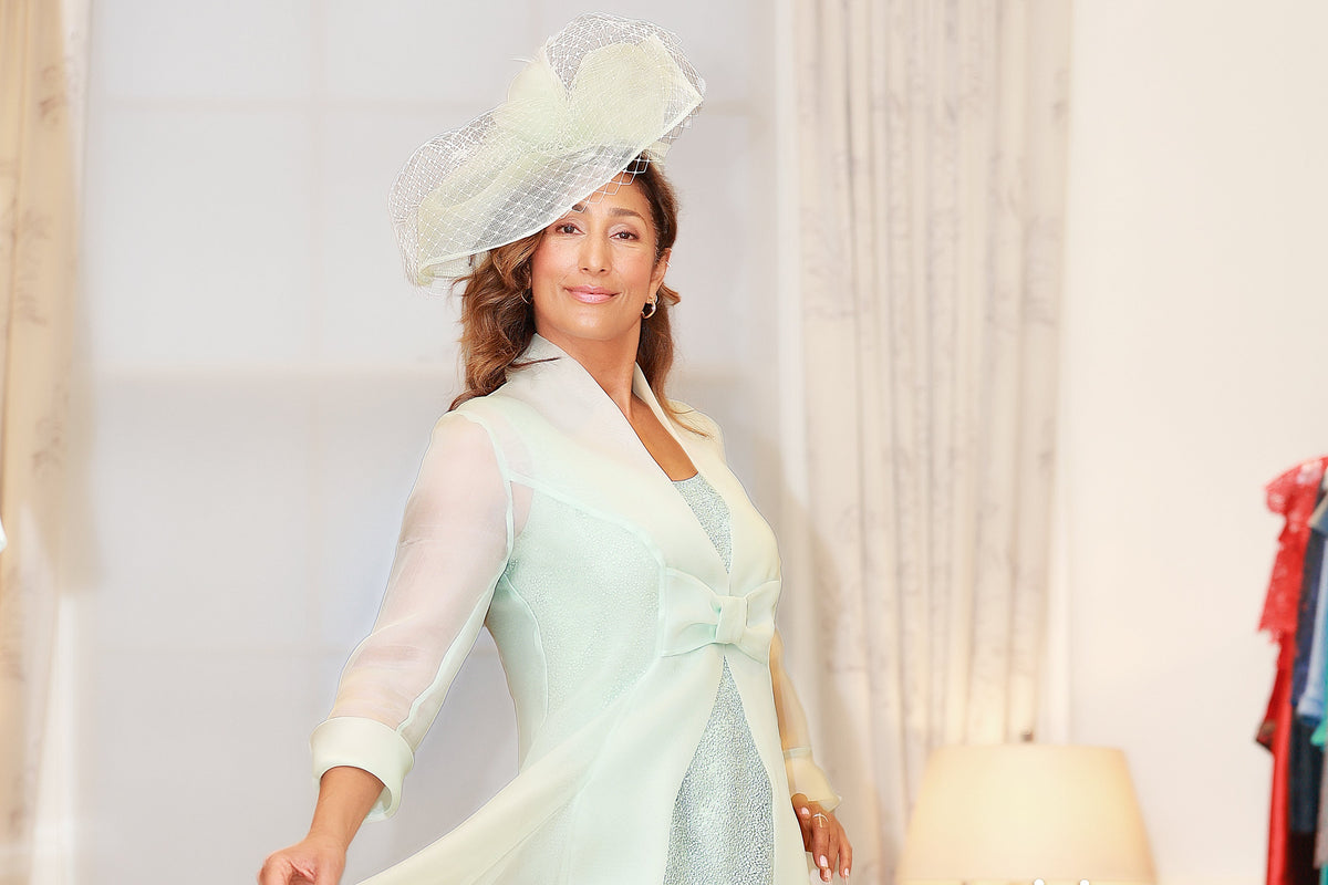 A woman modeling a light mint green midi dress with a matching sheer long-sleeve overlay, a large matching fascinator hat, and beige heels in an indoor boutique setting.