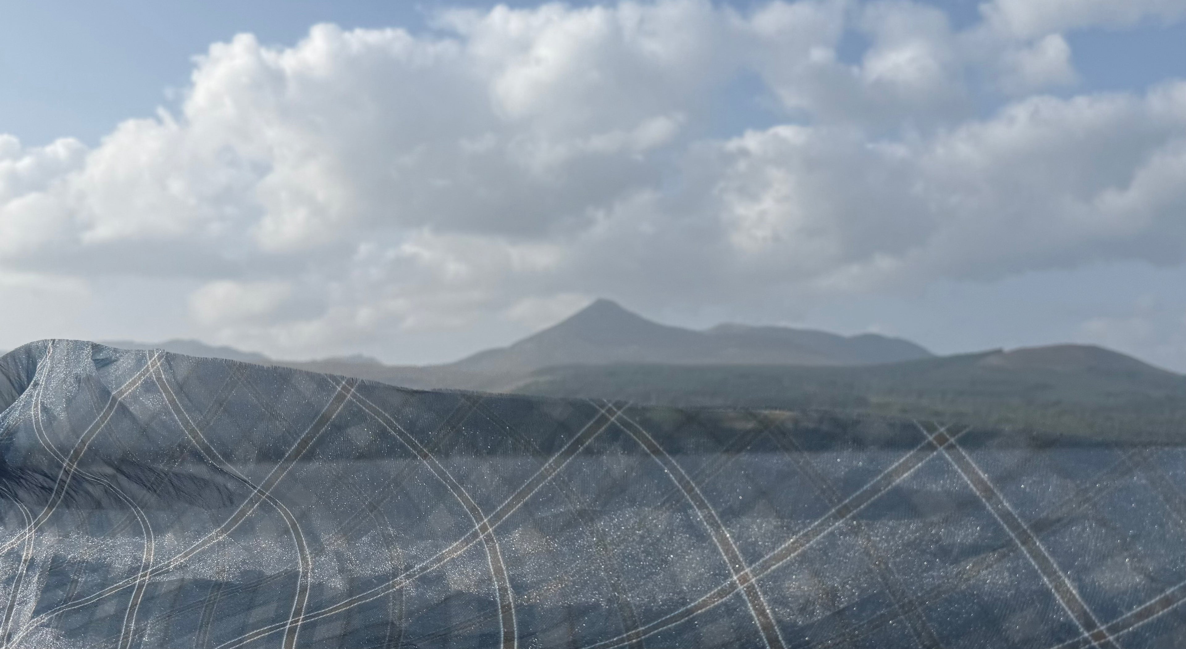 A close-up of a dark gray mesh fabric with a white diagonal grid pattern, draped over a surface with a mountain landscape and cloudy sky in the background.