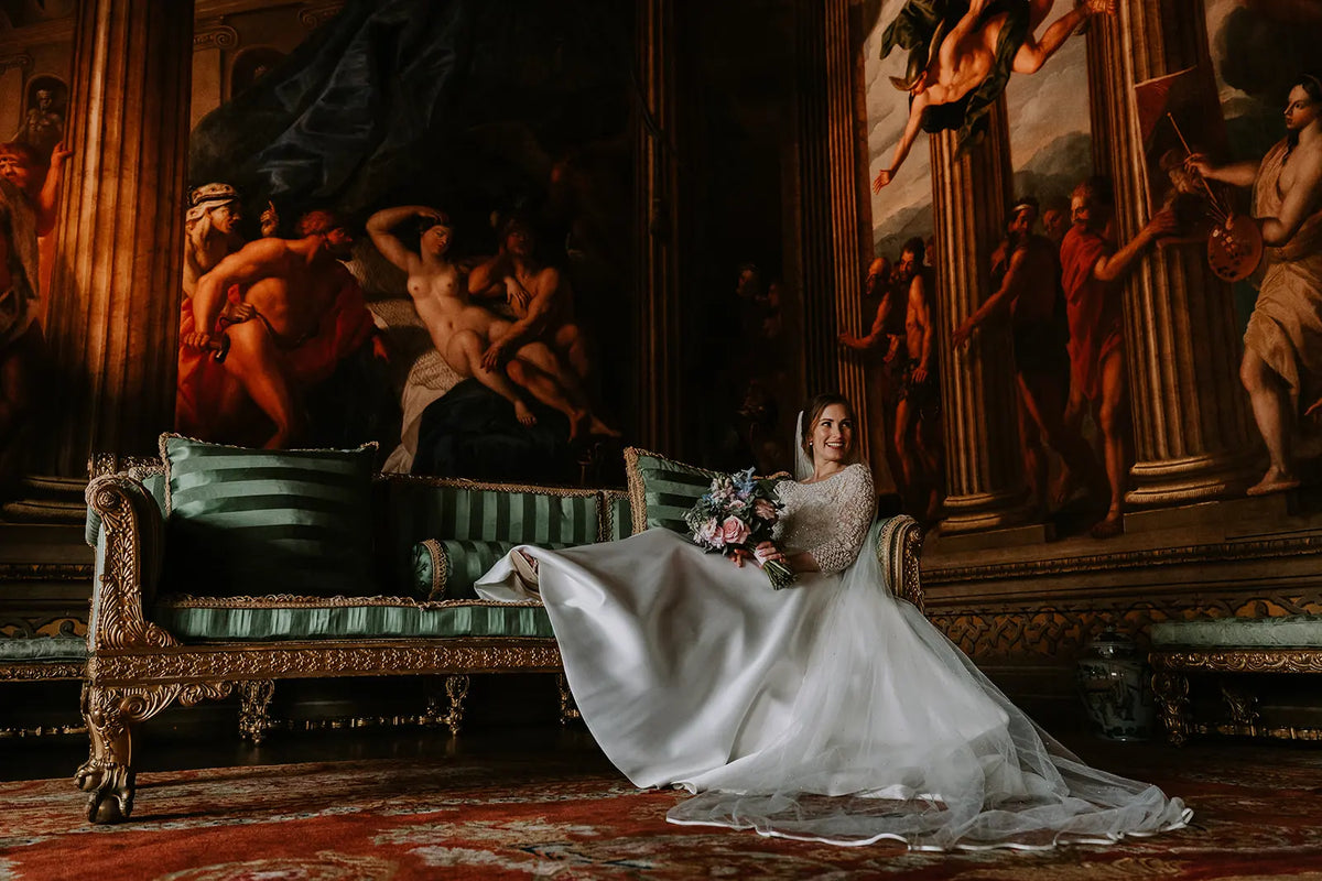 A bride in a long white lace wedding gown sits on an ornate green and gold sofa, holding a bouquet in a grand room with large classical paintings and warm ambient lighting.