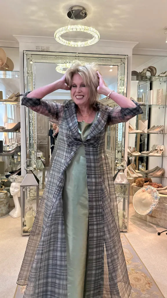 joanna lumley posing in a long gray plaid coat over a sage green satin dress inside a boutique showroom with mirrors and shoe displays.