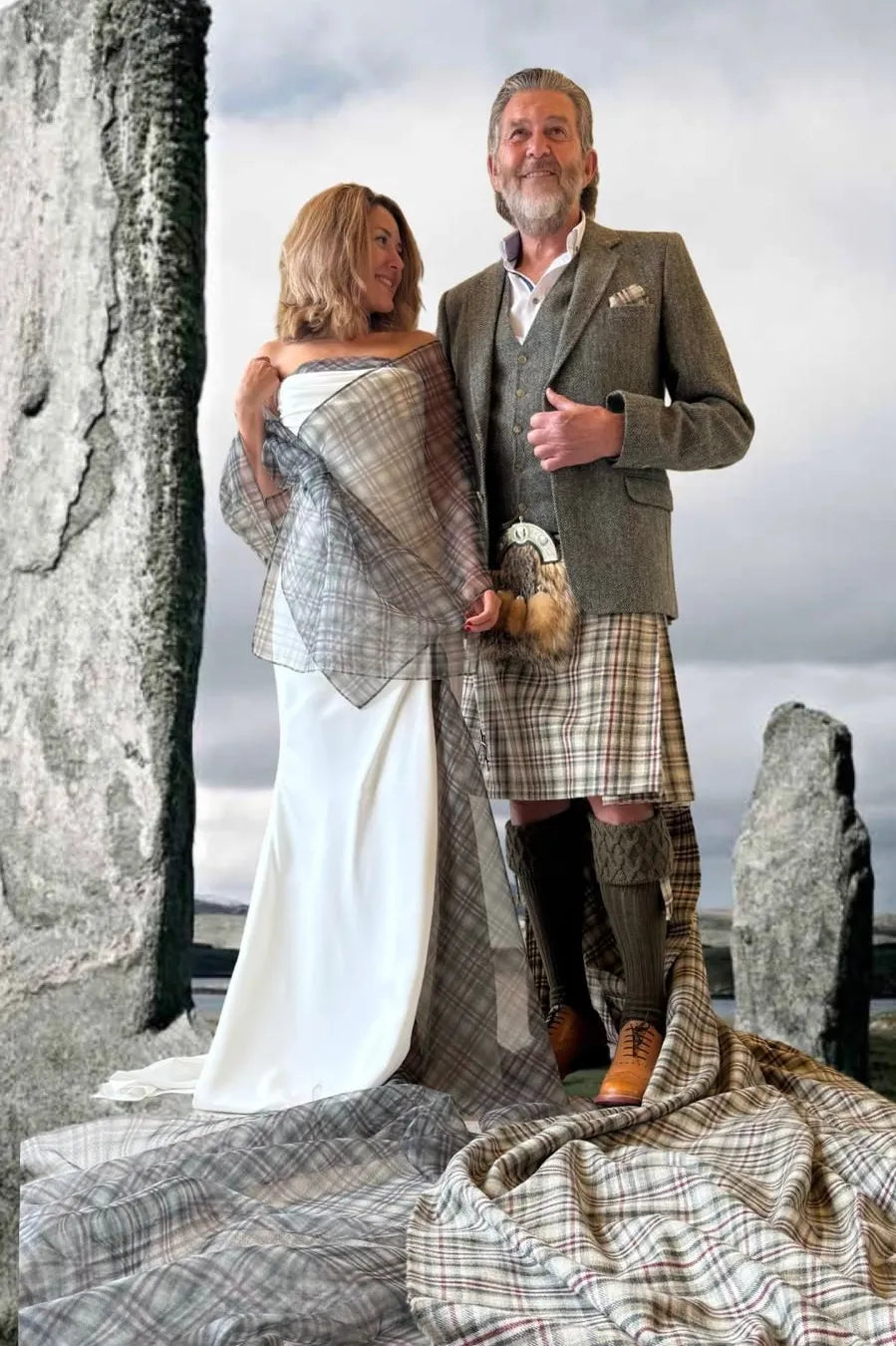 A man and woman posing outdoors in traditional Scottish attire, featuring a white wedding dress, a brown tweed kilt, and a plaid tartan wrap and blanket draped over the ground and around them, set against a cloudy sky and standing stones.