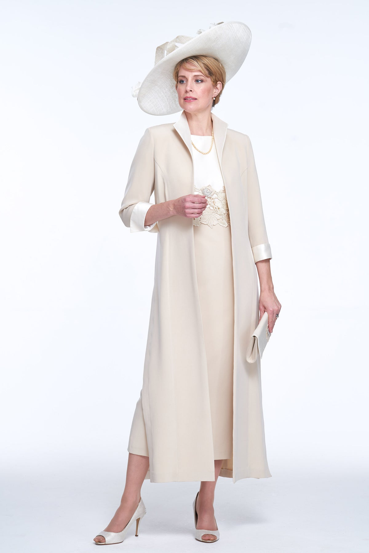 A woman modeling a cream-colored formal outfit consisting of a long open-front coat over a matching dress, a wide-brimmed hat, and white heels, photographed against a seamless white studio background.