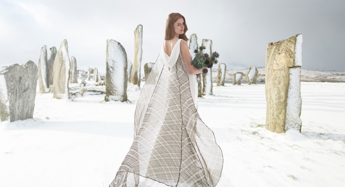 A woman stands in a snowy outdoor landscape with standing stones, wearing a long white sleeveless dress with a sheer beige-and-white plaid overlay and holding a bouquet of greenery.