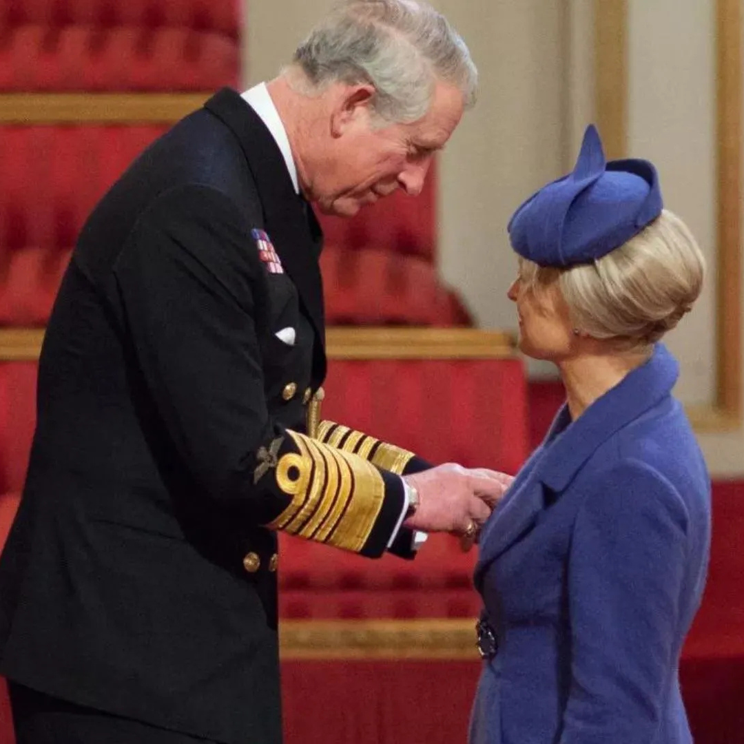Prince Charles in a dark navy military uniform with gold epaulets bows down his head to Queen Charlotte, who is wearing a blue coat and matching blue fascinator hat in a grand hall with red upholstered seating in the background.