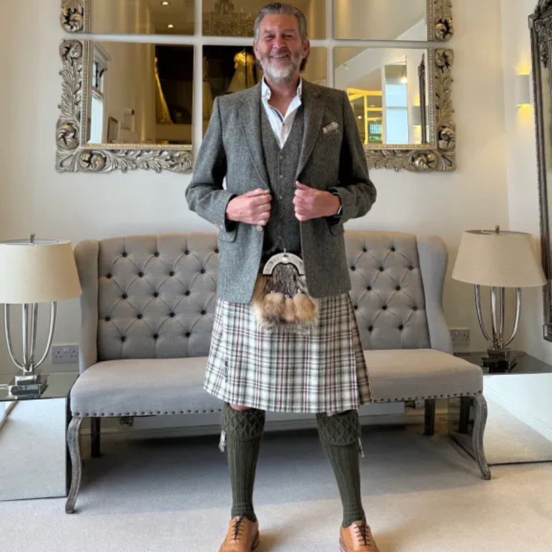 A man wearing a traditional Scottish outfit, including a gray tweed blazer, waistcoat, tartan kilt, green socks, and brown leather shoes, standing in front of a gray tufted bench and mirrors in an indoor setting.