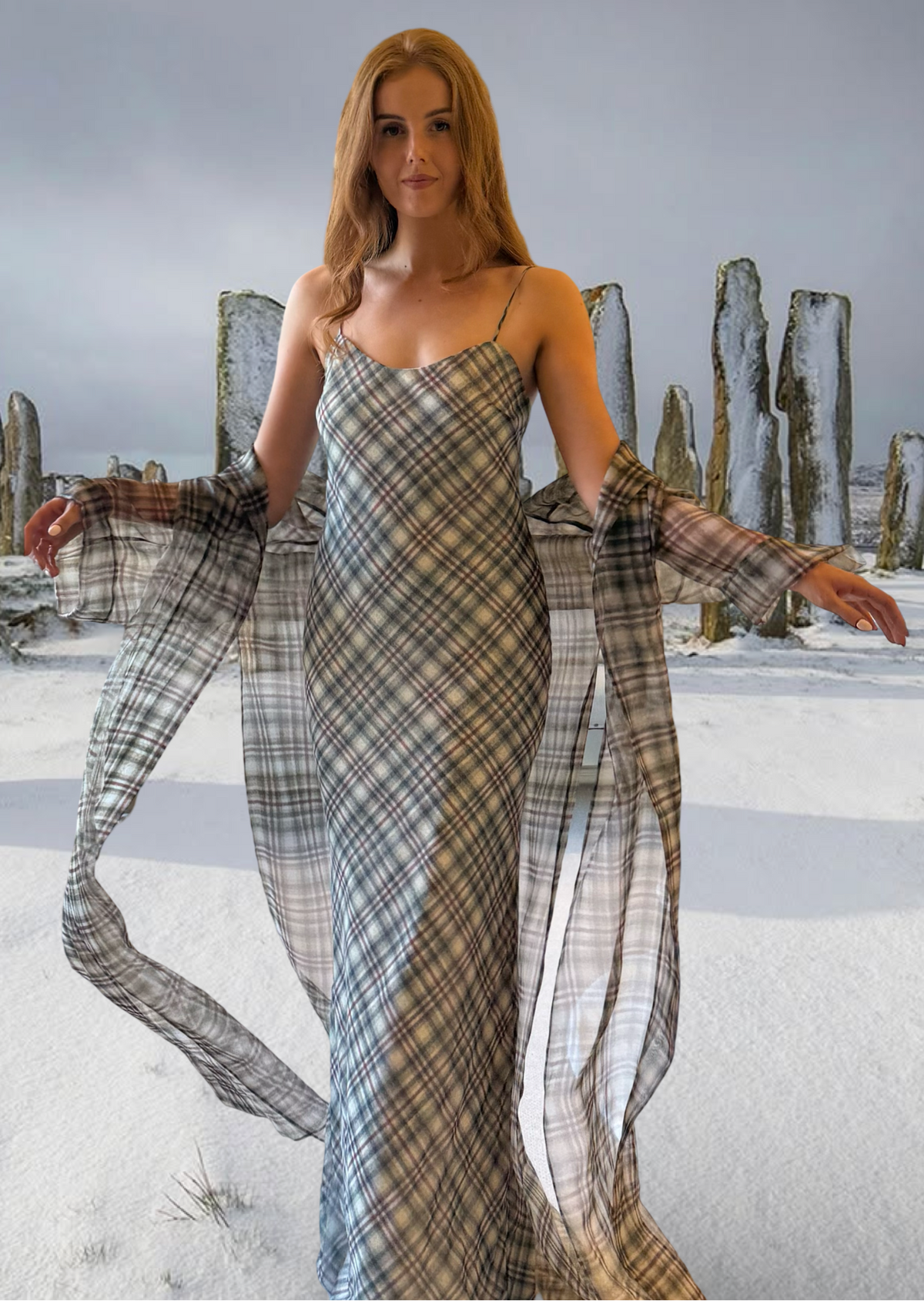 A woman stands outdoors in a snowy landscape wearing a long plaid slip dress with matching sheer draped sleeves. The background features large stone standing stones under an overcast sky.