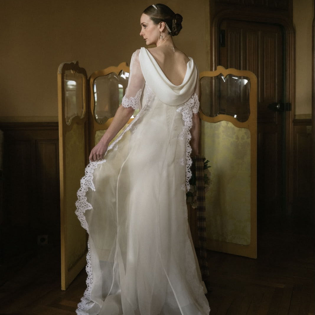 A woman wearing a flowing white wedding gown with sheer sleeves and lace trim stands indoors in a dimly lit room, holding out the train of the dress.