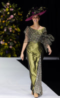A model walks a fashion runway wearing a floor-length olive green satin gown with a plaid draped overlay and a matching wide-brimmed purple fascinator hat decorated with greenery.
